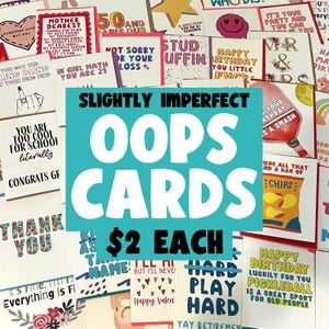 May include: A collection of greeting cards with various humorous and quirky messages. The cards are displayed on a table with a teal blue banner that reads "Slightly Imperfect Oops Cards $2 Each".