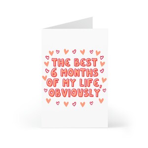 May include: A white card with pink hearts around the border. The text on the card reads "The best 6 months of my life, obviously."