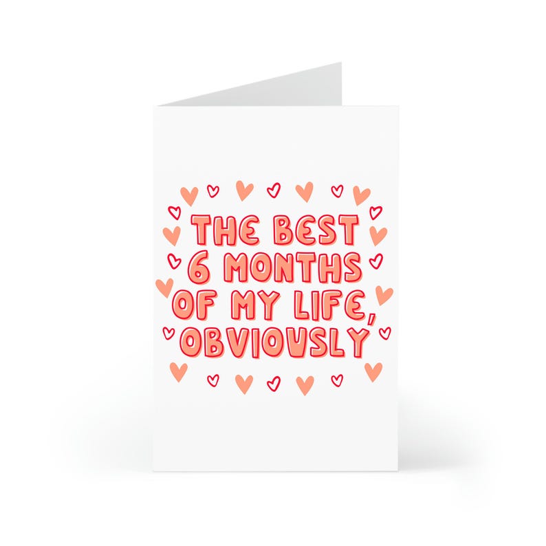 May include: A white card with pink hearts around the border. The text on the card reads "The best 6 months of my life, obviously."