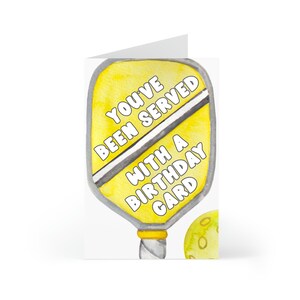 K&ouml;nnte beinhalten: Eine gelbe und wei&szlig;e Geburtstagskarte mit einer Aquarell-Illustration eines Pickleball-Schl&auml;gers und eines Balls. Der Schl&auml;ger tr&auml;gt den Text "You've Been Served With A Birthday Card".
