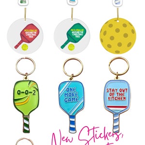 K&ouml;nnte beinhalten: Ein Set an Pickleball-Schl&uuml;sselanh&auml;ngern, Ornamenten und Aufklebern. Die Schl&uuml;sselanh&auml;nger zeigen einen Pickleball-Schl&auml;ger mit dem Text "0-0-2", "One More Game" und "Stay Out of the Kitchen". Es gibt auch einen gelben Pickleball-Schl&uuml;sselanh&auml;nger. Die Ornamente zeigen einen Pickleball-Schl&auml;ger mit dem Text "Pickleballin' Around the Christmas Tree". Die Aufkleber zeigen einen Pickleball-Schl&auml;ger mit dem Text "One More Game", "Stay Out of the Kitchen" und "0-0-2".