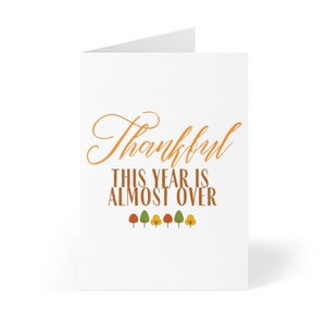May include: A white card with the text "Thankful This Year Is Almost Over" in orange. There are six small trees with orange, yellow, and green leaves at the bottom of the card.