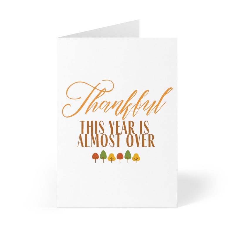 May include: A white card with the text "Thankful This Year Is Almost Over" in orange. There are six small trees with orange, yellow, and green leaves at the bottom of the card.