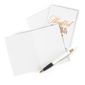 May include: A blank white card with a folded design. The card has a smaller card attached to the front with the text "Thankful the year is over" in gold lettering.