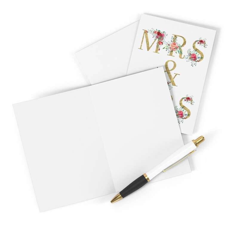 May include: A blank white card with a gold pen lying on top of it. The card is folded in half and has a smaller card on top of it. The smaller card has the words "MRS &" in gold letters with floral accents.