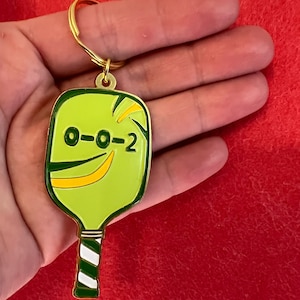 K&ouml;nnte beinhalten: Ein goldfarbener Schl&uuml;sselanh&auml;nger in Form eines gr&uuml;nen und gelben Pickleball-Schl&auml;gers mit einem wei&szlig;-gr&uuml;nen gestreiften Griff. Der Schl&auml;ger hat die Nummer "0-0-2" aufgedruckt.