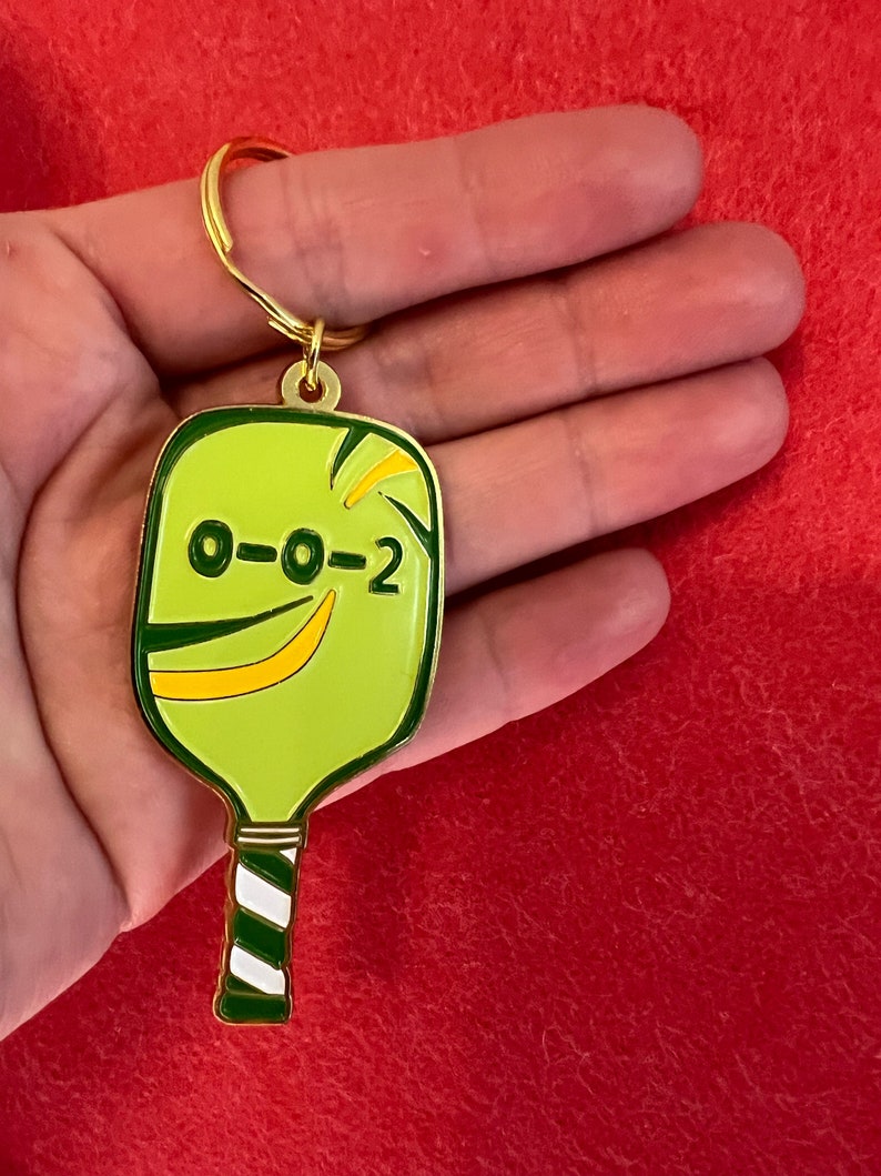 K&ouml;nnte beinhalten: Ein goldfarbener Schl&uuml;sselanh&auml;nger in Form eines gr&uuml;nen und gelben Pickleball-Schl&auml;gers mit einem wei&szlig;-gr&uuml;nen gestreiften Griff. Der Schl&auml;ger hat die Nummer "0-0-2" aufgedruckt.