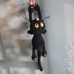 Könnte beinhalten: Eine handgefertigte schwarze Häkelkatze mit gelben Augen und weißen Akzenten. Die Katze hängt an einem orangefarbenen Ring. Die Katze hat lange Arme und Beine und einen langen Schwanz. Der Kopf der Katze hat einen weißen Punkt.