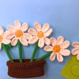 Op de afbeelding: Gehaakte madeliefjes in een bruine pot met groene stelen en gele centra. Een groene kaart met een hart ligt naast de bloemen. De achtergrond is lichtblauw.