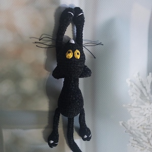 Peut inclure: Un jouet fait main en crochet noir représentant un chat avec des yeux jaunes et des détails blancs sur les pattes et les oreilles. Le chat est suspendu à l'envers, les bras levés. Le jouet a de longues moustaches et une petite tache blanche sur la tête.