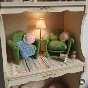 May include: Two miniature green crocheted armchairs with rounded backs, each featuring a small round cream-colored pillow. A light blue crocheted blanket rests on one chair. A tall thin lamp with a cream shade stands between the chairs on a striped rug.