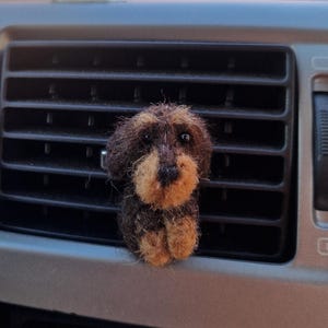 Könnte beinhalten: Eine kleine, nadelgefilzte Hundefigur mit dunkelbraunem und hellbraunem Fell, schwarzen Perlenaugen und einer schwarzen Nase. Die strukturierte, flauschige Skulptur besteht aus Wollfasern und zeigt einen sitzenden Hund mit Schlappohren und Schnauze.