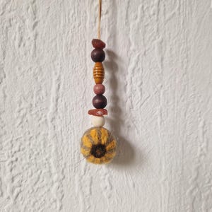 Könnte beinhalten: Ein hängendes Ornament mit einer vertikalen Schnur aus verschiedenen Perlen in Braun-, Beige- und Cremetönen, gekrönt von einem kleinen, unregelmäßigen bernsteinfarbenen Stück. Unten hängt eine runde, flauschige, gelb-braun gestreifte Filzkugel.