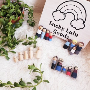 Pode incluir: Bonecas de madeira, algumas pintadas para representar famílias, estão dispostas sobre uma superfície branca. As bonecas têm rostos e roupas pintadas de forma simples. Uma caixa de madeira com um arco-íris e o texto "Lucky Lulu Goods" está no fundo.