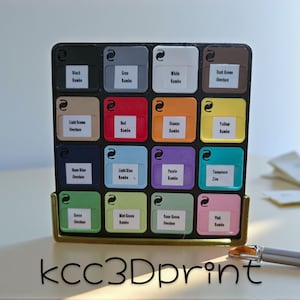 May include: A black and gold organizer with sixteen color swatches labeled with color names. The swatches are in various colors, including black, gray, white, red, orange, and green. A pen rests on the surface.