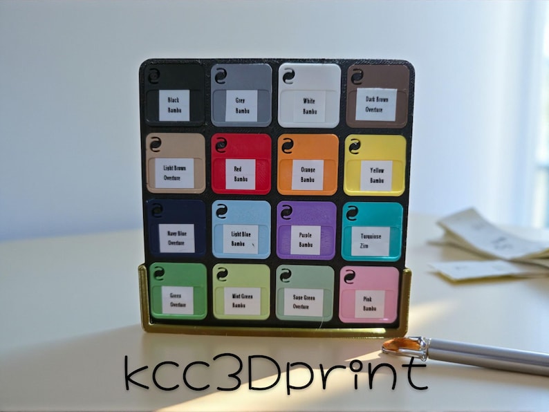 May include: A black and gold organizer with sixteen color swatches labeled with color names. The swatches are in various colors, including black, gray, white, red, orange, and green. A pen rests on the surface.