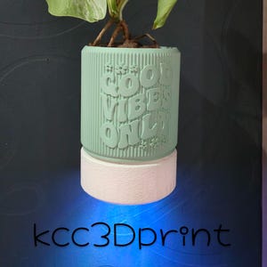 May include: A mint green planter with the words "GOOD VIBES ONLY" in a raised, stylized font. The planter sits atop a white base and holds a small plant. The text "kcc3Dprint" is at the bottom.