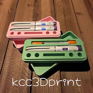 以下が含まれることがあります： インスリンペンを収納するピンクとグリーンの3Dプリントケース。ピンクのケースには2本のペン、グリーンのケースには1本のペンと注射器が入っています。「kcc3Dprint」の文字が下部に表示されています。