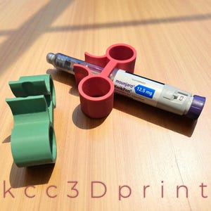 以下が含まれることがあります： 赤と緑の 3D プリントされたペンホルダーと、別の緑色のペンホルダー。 Mounjaro KwikPen 12.5 mg ペンが赤いホルダーに保持されています。 ペンは白、グレー、紫で、青い文字が書かれています。 テキスト「kcc3Dprint」が一番下にあります。