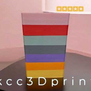 May include: A multi-colored, rectangular container with stacked layers of different colors, including pink, red, green, gray, purple, orange, and yellow. The text "kcc3Dprint" is at the bottom. The container is on a dark surface.