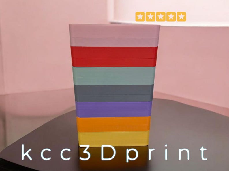 May include: A multi-colored, rectangular container with stacked layers of different colors, including pink, red, green, gray, purple, orange, and yellow. The text "kcc3Dprint" is at the bottom. The container is on a dark surface.