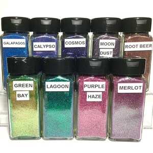 K&ouml;nnte beinhalten: Zehn kleine Glasbeh&auml;lter gef&uuml;llt mit buntem Glitter. Die Beh&auml;lter haben schwarze Deckel und Etiketten mit Namen wie "Galapagos", "Calypso", "Cosmos", "Mondstaub", "Wurzelbier", "Gr&uuml;ne Bucht", "Lagune", "Lila Nebel" und "Merlot".
