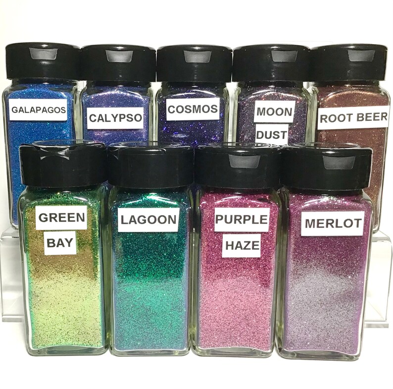 K&ouml;nnte beinhalten: Zehn kleine Glasbeh&auml;lter gef&uuml;llt mit buntem Glitter. Die Beh&auml;lter haben schwarze Deckel und Etiketten mit Namen wie "Galapagos", "Calypso", "Cosmos", "Mondstaub", "Wurzelbier", "Gr&uuml;ne Bucht", "Lagune", "Lila Nebel" und "Merlot".