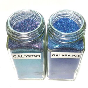 K&ouml;nnte beinhalten: Zwei Glasbeh&auml;lter gef&uuml;llt mit blauem Glitter. Der Beh&auml;lter links ist mit "CALYPSO" beschriftet und der Beh&auml;lter rechts mit "GALAPAGOS".