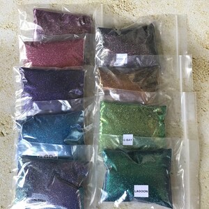 K&ouml;nnte beinhalten: Acht kleine Beutel mit schimmerndem Glitter, jeder mit einer anderen Farbe und einem anderen Namen. Die Farben sind Lila, Rosa, Blau, T&uuml;rkis, Gr&uuml;n und zwei Gr&uuml;nt&ouml;ne. Die Namen sind "Midnight", "Rose Gold", "Amethyst", "Lagoon", "Sea Glass", "Green Bay" und "Calpyso".
