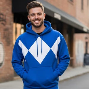 Pode incluir: Moletom azul com um padrão de losangos brancos no peito. O moletom tem um bolso frontal e um capuz. A pessoa está usando jeans pretos. O fundo inclui um edifício de tijolos e uma rua.