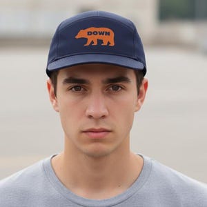 May include: A navy blue baseball cap with a flat brim. The cap features the word "DOWN" above an orange bear silhouette. The cap is worn by a person in a grey t-shirt.