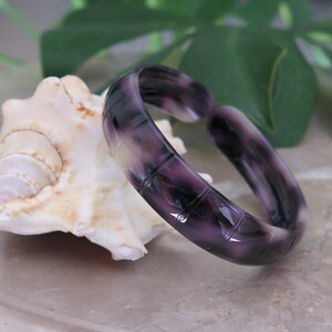 May include: A purple and white marbled bangle bracelet rests on a seashell. The bracelet is made of a translucent material with a square pattern. The background includes a green leaf and a light-colored surface.
