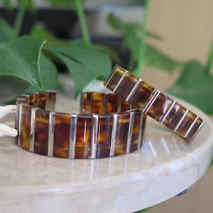 Può includere: Un set di due braccialetti abbinati. Uno è un braccialetto a polsino e l'altro è un braccialetto rettangolare più piccolo. Entrambi presentano un motivo a scaglie di tartaruga con dettagli in metallo color argento. I braccialetti sono esposti su una superficie bianca con sfondo di fogliame verde.