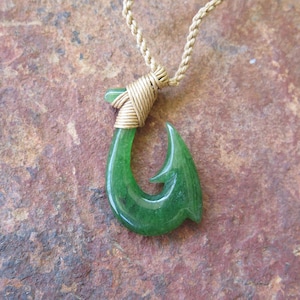 Puede incluir: Un collar con un colgante de gancho de jade verde y un envoltorio de alambre dorado. El gancho es un símbolo tradicional hawaiano de buena suerte y prosperidad.