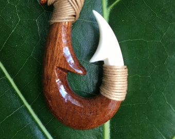 Puede incluir: Un colgante de anzuelo de pescado de madera marrón con una punta de hueso blanco, colgando de un collar de cordón marrón. El colgante está sobre un fondo de hoja verde.