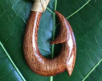May include: A brown wooden fish hook necklace with a tan cord. The hook is polished and has a natural wood grain. The necklace is on a green leaf.