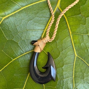 May include: A black pendant necklace featuring a curved hook design. The hook is suspended from a braided tan cord. The necklace is displayed on a large green leaf.