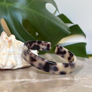 May include: A tortoiseshell-patterned cuff bracelet. The open-ended bangle features a cream-colored base with dark brown spots. The bracelet is displayed on a marble surface, with a seashell and a large green leaf in the background.