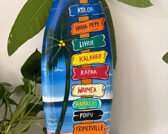 May include: A wooden surfboard painted with a blue sky, palm trees, and a beach scene. The surfboard has a series of painted signs pointing to different locations on the island of Kauai, Hawaii: Koloa, Hana Pepe, Lihue, Kalaheo, Kapaa, Waimea, Hanalei, Poipu, Princeville, Wailua Falls, and Kauai.