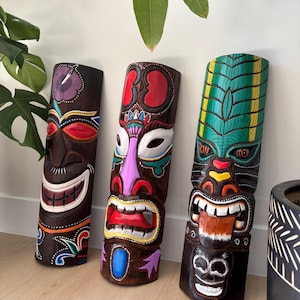 以下が含まれることがあります： カラフルなペイントのディテールが施された木製のティキマスクが3つ。マスクはそれぞれ表情とデザインが異なります。マスクは茶色で、赤、緑、青、黄、白、黒のペイントが施されています。