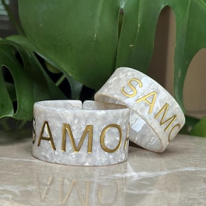 May include: Two wide cuff bracelets made of white pearlescent material with a marbled texture. Each bracelet features the gold-coloured text SAMOA embossed on the front. The bracelets have an open-back design and a smooth, glossy finish.