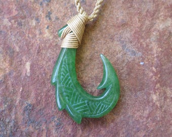 Puede incluir: Un colgante de anzuelo de pescado de jade verde en un collar de cordón marrón. El anzuelo está tallado con un diseño tradicional y tiene una envoltura de alambre dorado en la parte superior.