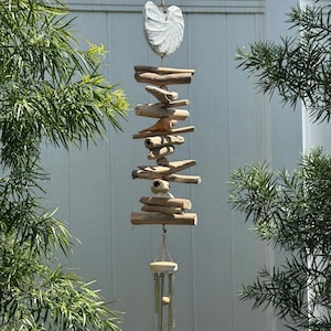 May include: A handcrafted wind chime with a white, shell-shaped top and a series of natural wood pieces suspended below. The chime features several metal tubes that hang from a wooden base, creating a soothing sound. The wind chime is hanging outdoors.