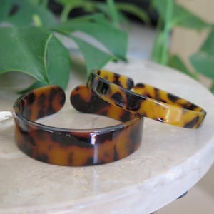 Può includere: Tre braccialetti in tartaruga sono esposti su una superficie di marmo. I braccialetti hanno una finitura lucida e un motivo di tonalità marrone scuro e ambra. Il braccialetto aperto è accanto a due braccialetti chiusi. I gioielli sono un accessorio di moda.