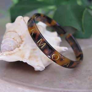 Può includere: Un braccialetto a cerchio con motivo tartarugato con lettere dorate che compongono la parola "SAMOA". Il braccialetto aperto è appoggiato su una conchiglia bianca, con fogliame verde sullo sfondo. Il braccialetto è di colore marrone caldo.