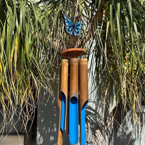 Puede incluir: Un carillón de viento de bambú con un acento de mariposa azul. El carillón de viento presenta tubos de bambú natural con interiores pintados de azul, suspendidos de un disco de madera. La mariposa es azul y negra.