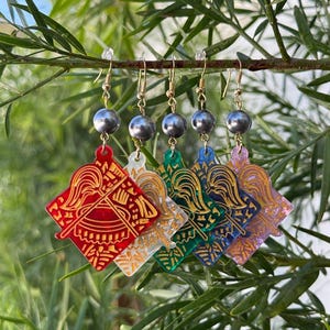 Puede incluir: Cinco pendientes colgantes con colgantes cuadrados de acrílico en rojo, blanco, verde, azul y rosa. Cada colgante muestra un diseño grabado en color dorado de un casco alado. Una perla gris oscuro se sitúa sobre cada colgante.