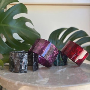 May include: A collection of colorful, patterned napkin rings. The rings are in shades of black, purple, green, and red. They are arranged on a round, marble-like surface, with large green leaves in the background.