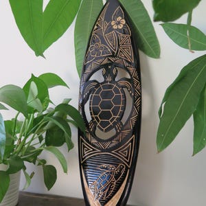 Puede incluir: Decoración de pared de madera negra en forma de tabla de surf con un diseño tallado de una tortuga y flores. La tortuga está en el centro del diseño y está rodeada de un patrón geométrico. La tabla de surf mide aproximadamente 50 cm de largo.