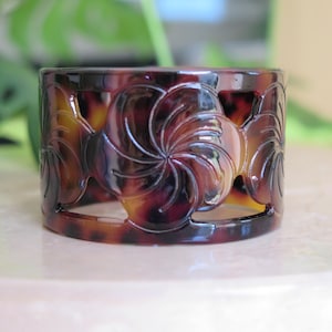 May include: A decorative cuff bracelet with a tortoiseshell pattern. The bracelet features a series of cut-out floral designs. The colors are shades of brown and amber. The bracelet is approximately 1.5 inches wide.
